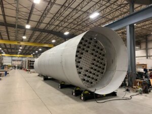 A large cylindrical stainless steel structure with precision-laser cut holes, showcased in a spacious manufacturing facility demonstrating advanced metal fabrication techniques and precision engineering.