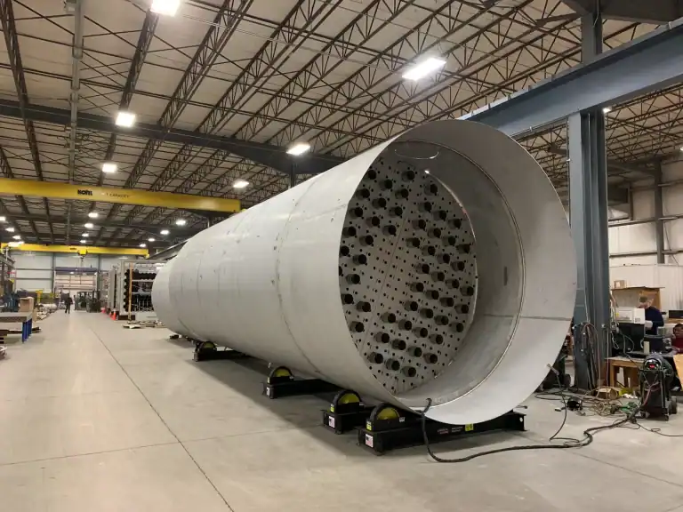 A large cylindrical stainless steel structure with precision-laser cut holes, showcased in a spacious manufacturing facility demonstrating advanced metal fabrication techniques and precision engineering.