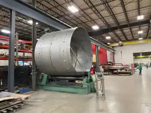 A spacious industrial facility showcasing stainless steel fabrication with a focus on custom precision metal fabrication. A large cylindrical metal structure is being worked on a rolling machine, surrounded by tools and workers in a collaborative manufacturing environment.